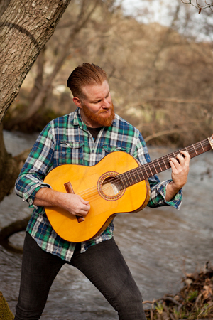 Hipster man with red beard playing a guitar in the field