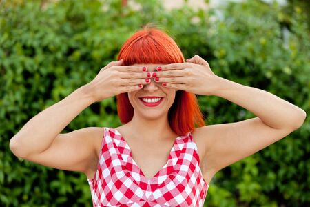 Red haired woman with red checkered dress covering her eyes in a park