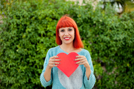 Relaxed red haired woman with blue jersey in the park holding a red heart
