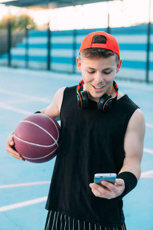 Attractive guy basketball player with a ball looks at his mobile phone and smiles while taking a breakの写真素材