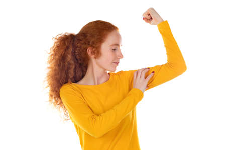 Indoor portrait of young redhead female with wavy long hair positively shows her muscles and smile,...