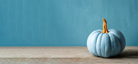 Single blue pumpkin with orange stem on rustic wooden table against textured blue background in minimalist autumn concept by Generative AIの素材