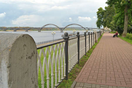 Volga river embankment in Rybinsk. View from the park to the bridge and cloudy sky on the horizon. Bench, paved path, in the foreground a lattice fence.の写真素材
