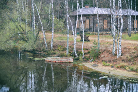 The shore with an overturned old rowing boat and an old wooden house among the trees in autumn...