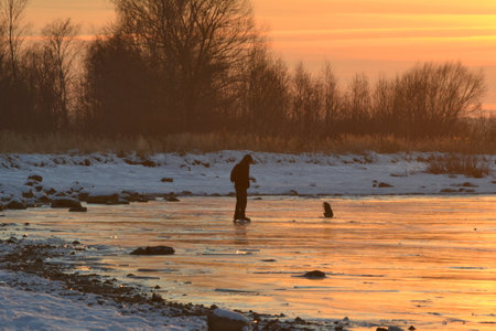 Golden sunset. Young adult man's silhouette on skates standing at sunset in dusk on frozen lake ice. Enjoying sport in cold weather. Outdoor activities on weekends.の写真素材