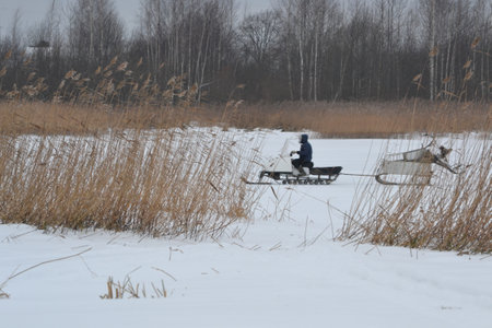 Fisherman on a snowmobile. Dry reed on lake shore on foreground, black trees and houses in the back. Gray gloomy winter landscape in inclement weatherの写真素材