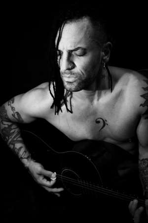 Man playing guitar and singing shirtless. Dark background