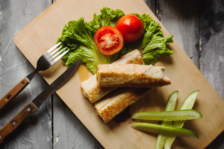 Grilled Kebab on a wooden board with tomatos, lettuce, and cucumber. Presentation with fork and serrated knife. Top view angleの写真素材