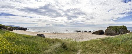 Panoramic view of beach looking out to sea on the West coast of Scotland surrounded by rocks,grass...