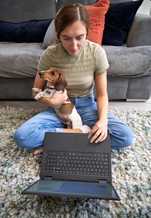 Young woman sitting on floor in front of sofa at home working on laptop computer with pet dog on her...