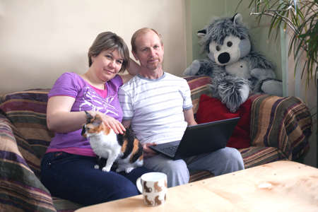 A middle-aged married couple siting at home with a laptop. On the sofa with catの写真素材