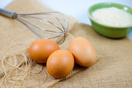 Chicken eggs on sack with whisk and a bowl of rice background (selective focus)の写真素材