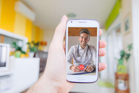 Hand holding mobile phone having video call with doctor on the screen. Videoconference at home with...