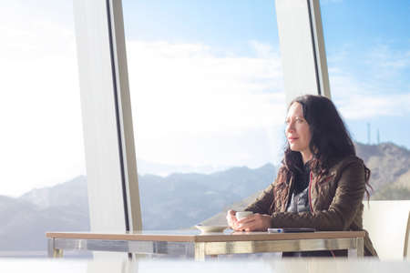 Young woman sitting alone in cafe looking trough big windows. Mountains and blue sky outdoors in...