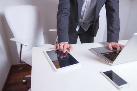 Front view of businessman working on laptop and tablet while standing at home officeの写真素材