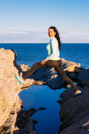 Sporty beautiful woman standing on the sea rocks and looking at camera. Blue ocean and sky in the backgroundの写真素材