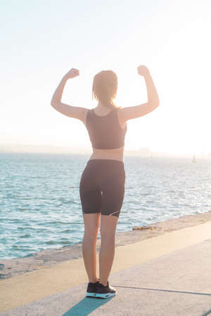 Back view of sports girl with arms raised standing outdoors near the sea feeling strongの写真素材