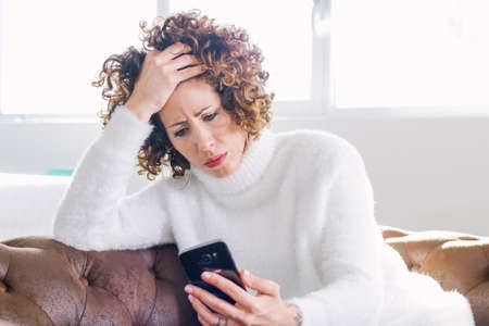 Worried woman checking smartphone sitting on a couch at home