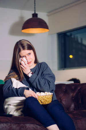 Girl at home sitting on the sofa with a popcorn bowl and crying because she is watching a sad movie...