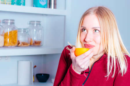 Portrait of beautiful young woman in the kitchen smelling an orange freshly cut in half. Healthy food concept.の写真素材