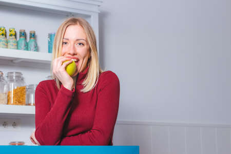 Portrait of happy young woman biting a green apple in the kitchen. Healthy food and lifestyle...
