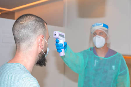Healthcare worker wearing protective face shield measuring customer temperature with digital...