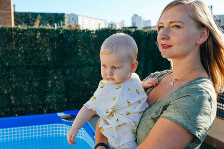 Portrait of young woman holding her baby daughter in her arms outdoors. Mother and baby on a terrace with swimming pool in the backgroundの写真素材