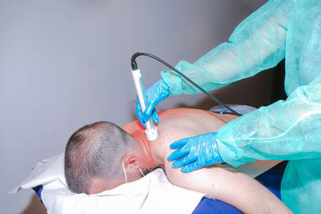 Physiotherapist using a laser device on the patient's back. Close up view of hands with latex gloves applying a muscle treatment to man lying on a stretcherの写真素材