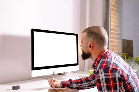 Young man in front of computer with blank screen taking notes at home office desk.の写真素材