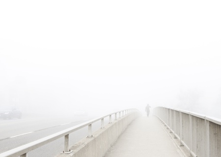 Solitary man in distance walks away, disappearing into the fog. Pedestrian walkway on bridge recedes in perspective toward subject at vanishing point. High key, desaturated, horizontal with copy space.の写真素材