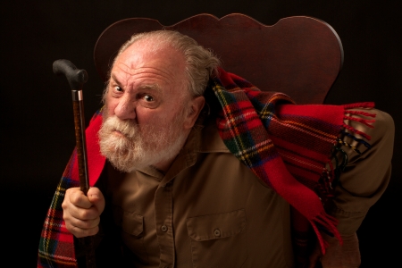 Leaning forward in his chair, an old man with gray beard looks directly at the camera, frowns, raises his cane and shakes it  He has a dark tan shirt and red plaid wool shawl  Horizontal composition with spot lighting and a black background の写真素材