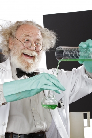 Cheerful mad senior scientist in lab measures green liquid in beaker  Frizzy grey hair, round glasses, lab coat, aqua rubber gloves, blank blackboard, vertical, high key, copy space の写真素材