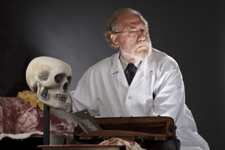 Evil doctor with surgical tools and bloody corpse  Sinister expression, dark background with dramatic low angle spot lighting  Horizontal, copy space の写真素材