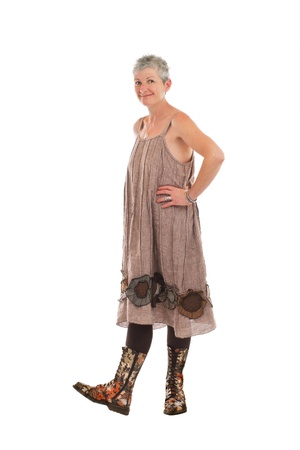 Cheerful friendly older woman stands in flowered boots and brown cotton shift dress. She has short gray hair. Isolated on white background, vertical.の写真素材