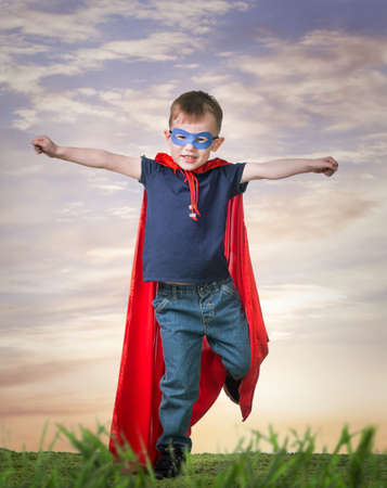 A boy in a Superhero costume stands on the grass in a red cloakの写真素材