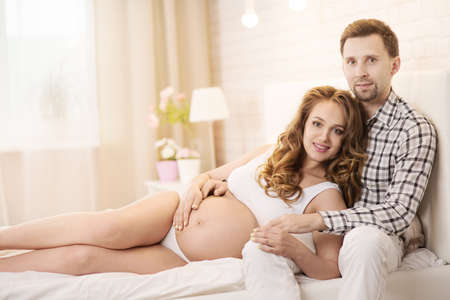 Pregnant wife and husband are sitting on the bedの写真素材