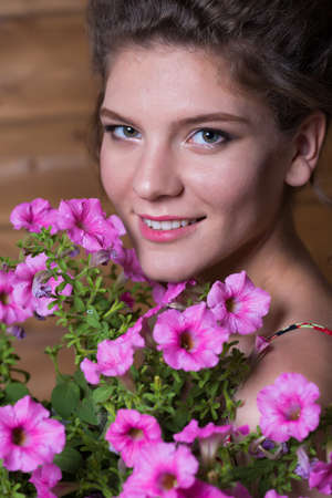 Beautiful young woman dressed in a rustic dress holding a pot of ampel Petunia flowers against a wooden wall backgroundの写真素材