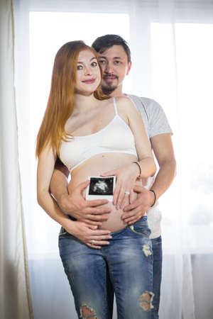 Husband and pregnant wife in a room with a window, happy couple 