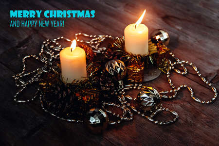 Lighted candles in candle holders with pine cones, tinsel, decorative toy balls and garland on wooden background in dark colors. Merry Christmas Greeting Card wallpaper. New Year winter decor concept.の写真素材