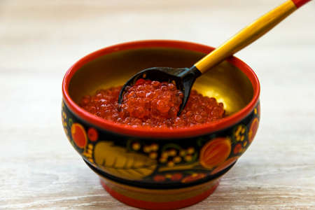 Fresh red caviar in wooden painted traditional style Khokhloma bowl with spoon on rustic bleached white wood table background. Close up. Selective focus. Copy spaceの写真素材