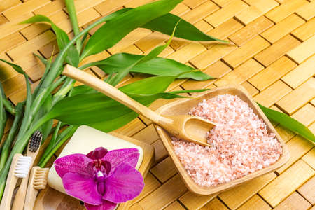 Spa, zen, massage concept. Pink himalayan bath salt with spoon, bamboo leaves, purple orchid, soap in soap dish on wooden carpet background. Top view. Close up. Selective focus. Text copy space.の写真素材