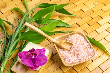 Spa, zen, massage concept. Pink himalayan bath salt with spoon, bamboo leaves, purple orchid, soap in soap dish on wooden carpet background. Top view. Close up. Selective focus. Text copy space.の写真素材