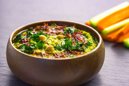 Homemade chickpea green hummus with herbs in round bamboo bowl, celery and carrot stick on black slate background. Close up. Selective focus. Copy spaceの写真素材