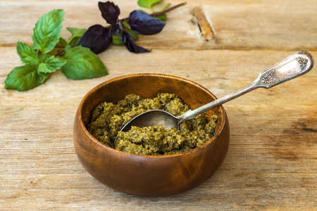 Basil pesto in bamboo bowl with metal spoon and green purple leaves on wooden table backgroundの写真素材