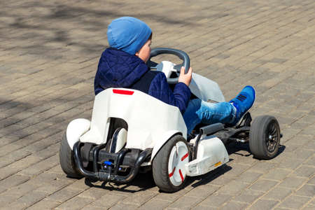 Kid boy back view blue jacket, hat, jeans driving white children pedal go-kart in park springtime.