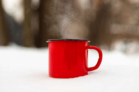 Red enamel mug cup coffee hot drink in snow against winter forest background. Close up. Copy spaceの写真素材