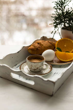 Coffee cup, croissant, egg, fresh orange fruit on white wooden tray against blur window backgroundの写真素材