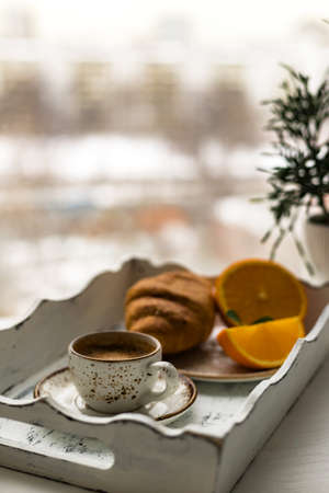 Coffee cup, croissant, fresh orange fruit on white wooden tray against blur window backgroundの写真素材