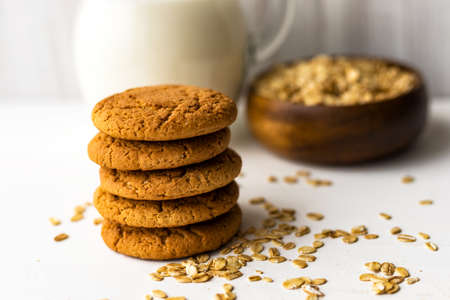 Heap oatmeal cookies and oat cereal flakes bowl on white table on milk glass jug backgroundの写真素材