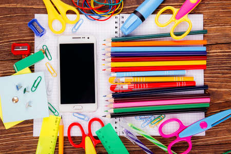 Open blank notebook, white smartphone with frame of school supplies on a wooden table background....